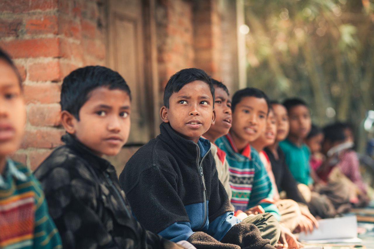 why-choose-us A group of children from Bangladesh sitting outdoors, engaged in learning at a rural school.